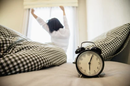 Schwarzer Wecker auf dem Bett, im Hintergrund streckt sich eine Person am Morgen – flexible Gastarife der EV Greiz für Vielverbraucher außerhalb des Netzgebietes