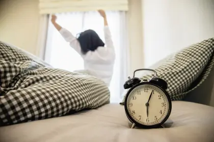Schwarzer Wecker auf dem Bett, im Hintergrund streckt sich eine Person am Morgen – flexible Gastarife der EV Greiz für Vielverbraucher außerhalb des Netzgebietes