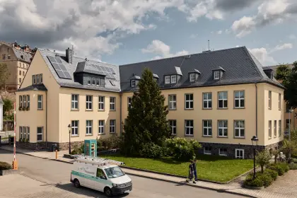 Das Gebäude der EV Greiz im Sonnenschein. Im Vordergrund fährt ein Auto und zwei Männer laufen durchs Bild in Handwerkerklamotten.